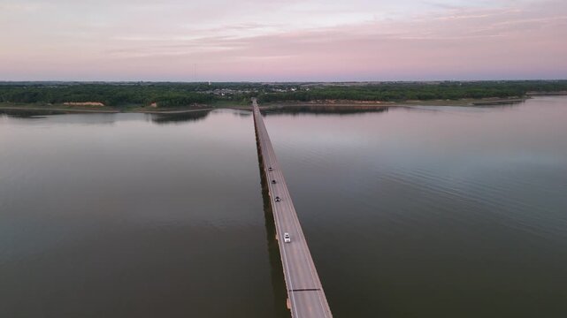 Aerial view of cars driving across long bridge over calm river at sunset.
