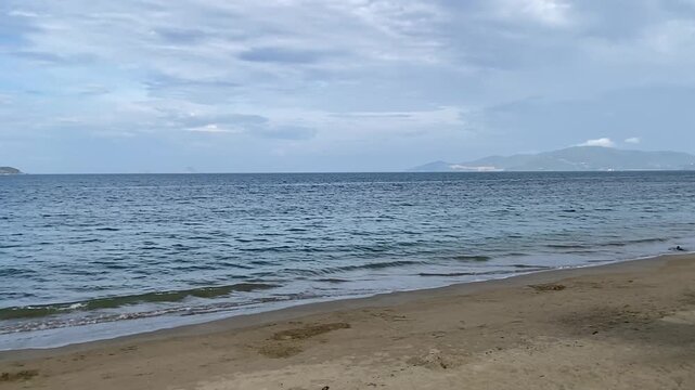 Calm sea waves on the sandy beach of North Nha Trang in Vietnam, beautiful coastal landscape of Ba Lang area with mountain islands on the horizon under cloudy sky.