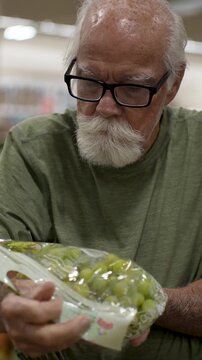 An elderly man browses through grapes at a grocery store to choose fresh produce. He inspects the packaging before deciding.