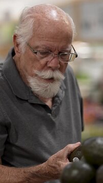 An elderly man examines avocados while shopping in a grocery store. He focuses on selecting fresh produce.