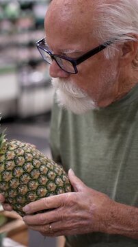 An elderly man looks closely at a pineapple while shopping in a grocery store in the afternoon. He appears focused on his task.