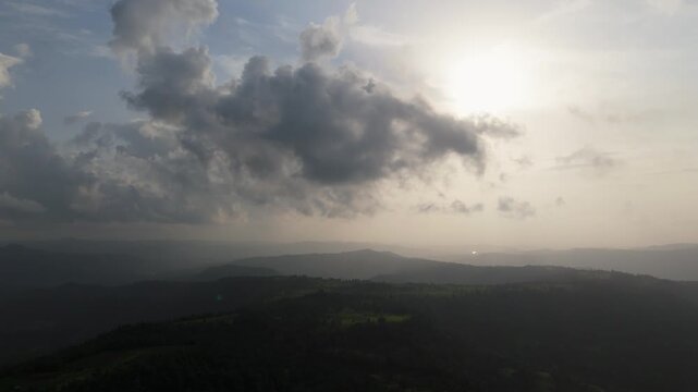 Aerial drone footage capturing a hazy sunset over the Western Ghats (Sahyadri mountain range), India. The hazy sun glows through layered clouds above rolling, forest-covered hills