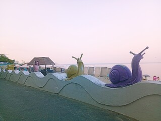 Large colorful decorative snail sculptures in purple, yellow, and pink colors crawling on a wavy concrete promenade border next to a sandy beach and calm sea during golden hour. © Oksana