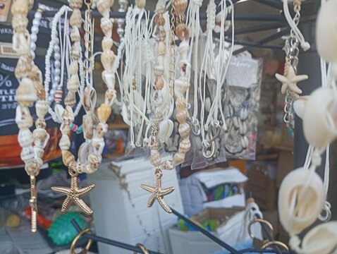 Close-up of traditional seaside souvenir necklaces and bracelets made from natural seashells, cowrie shells, and golden starfish pendants hanging on a display rack at a beach market.