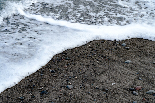 Gentle white ocean wave foam washing over dark volcanic sand beach, natural coastal tide line background.