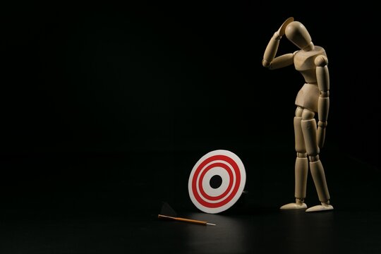 Wooden mannequin looking down at a missed target and fallen dart, symbolizing failure, mistake, and disappointment. Conceptual business metaphor on black background.