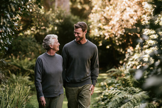 Adult son walking through a lush garden with his elderly mother, sharing laughter and companionship in a warm scene of family connection, support and active aging.
