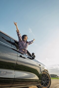 A woman with long hair joyfully leans from the window of a black car, raising her arms in celebration. She is surrounded by a picturesque landscape under a bright blue sky during daytime