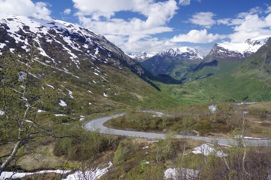 Road leads through spring-like landscape past partly snow-covered mountains downhill, alpine meadows, Langvatn, Geiranger, Norway