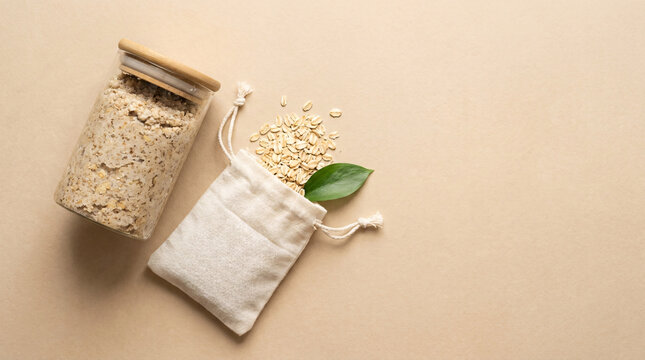 Rolled oats spilling from a drawstring pouch next to a glass jar filled with oats, set on a textured beige background with a green leaf, natural food ingredient and healthy breakfast concept