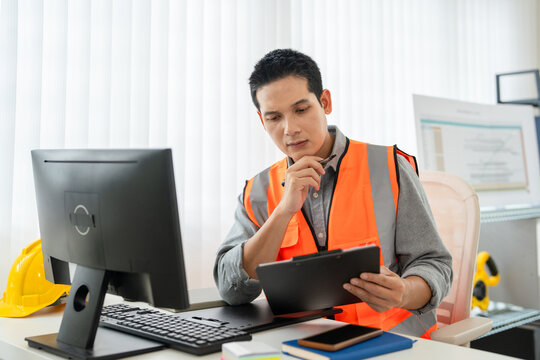 A young foreman or civil engineer in safety helmet and Reflective Safety Vest working at a desk in construction site office with Construction blueprint drawings plans and laptop.
