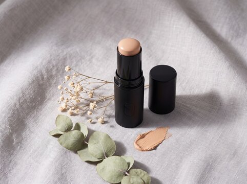 A foundation stick with a creamy swatch on a textured linen fabric background, decorated with dried flowers and eucalyptus leaves.