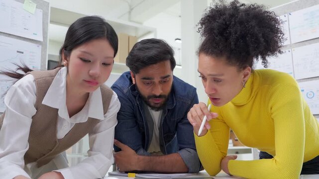 Three colleagues lean over a desk covered in printed charts and a tablet, closely reviewing data together during a focused analysis session in a modern office lined with project boards.