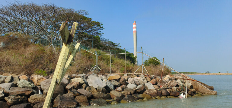 A natural landscape featuring a breakwater and industrial chimneys in the background.