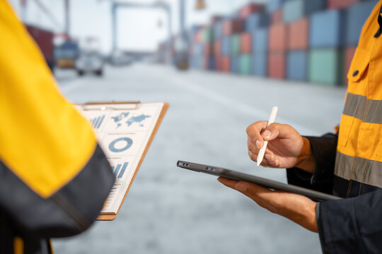 Two port workers in high visibility jackets review cargo documents and sign on a tablet beside stacked shipping containers at a busy freight terminal.