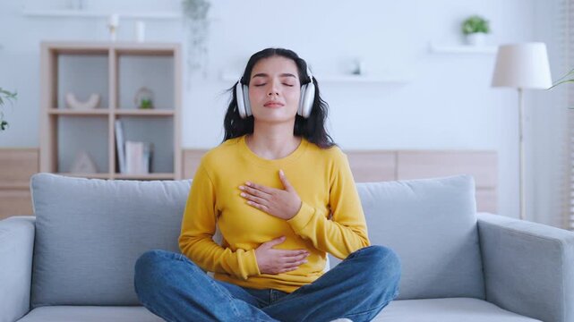 Asian woman meditates and practices Zen meditation at home, focusing on breathing exercises and seeking spiritual peace in her living room. She sits on the sofa, listening podcast to headphones