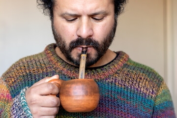Close-up of a focused South American man enjoying a cup of yerba mate inside his home while he...