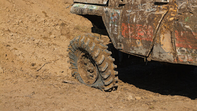 Close up of rugged off road tire caked in brown mud on dirt track. Vehicle body splattered with mud, showing racing wear. Warm sunlight highlights texture, creating dramatic shadows
