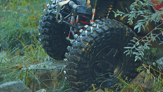 Close up of rugged off road vehicle tires caked in mud navigating rocky terrain. Green foliage frames the shot. Natural daylight highlights tire tread suspension