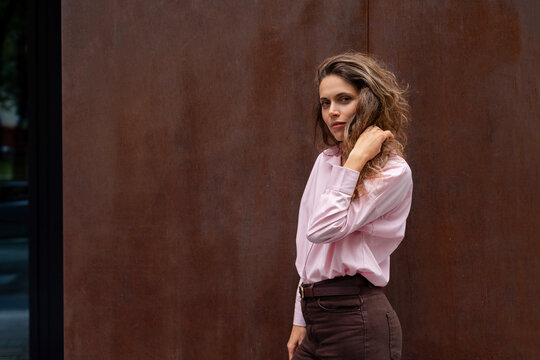 Young woman touching her curly hair against brown wall looking at camera