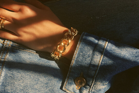 Close-up of a woman's hand wearing a chunky gold chain bracelet and ring, adorned with sunlight and denim texture