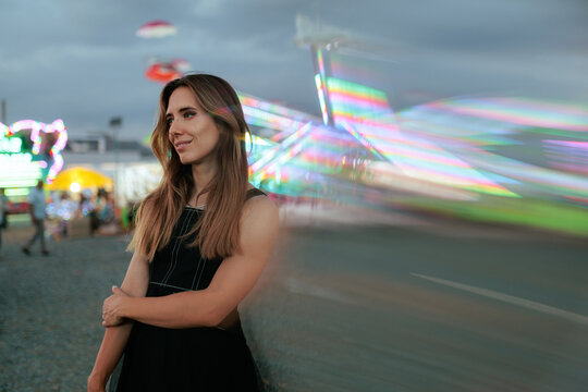 Attractive Young Woman Posing At Carnival Amusement Park At Night. Smiling lady stands near colorful blurred moving carousel lights in the evening
