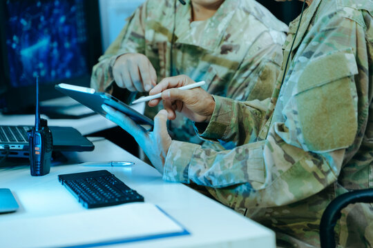 Two military officers in camouflage uniforms analyze tactical cybersecurity data on a tablet and multi-monitor displays