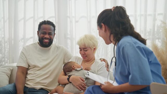 Nurse in blue scrubs reviews her notes and talks through newborn care advice with a multiracial couple holding their baby on the living room sofa during a home visit.