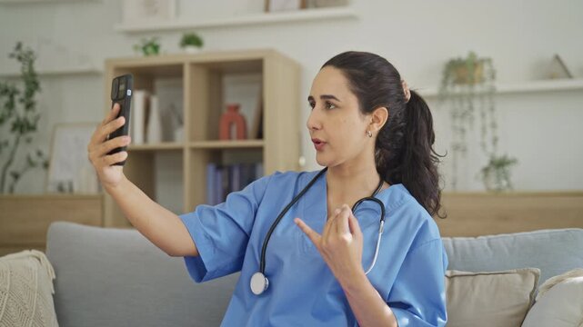 Nurse in a blue surgical scrubs raises her smartphone for a remote video call, using gestures to explain something to a colleague in a brightly lit living room.