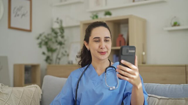 Young nurse in blue scrubs gestures while speaking during a telehealth video call on her smartphone, working remotely from a living room.