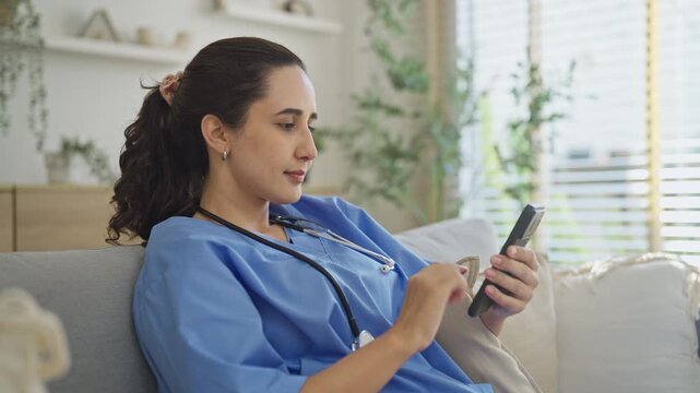 Smiling nurse in blue scrubs leans back on a comfortable sofa and scrolls her smartphone during a short break between home health visits in a bright living room.