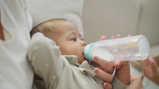 Close view of a newborn baby held in a parent's arms drinking formula milk from a bottle, cared for gently at home in soft natural light on the living room sofa.