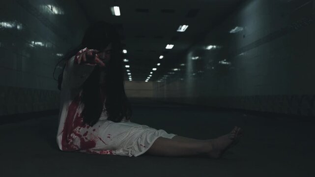Scary halloween ghost in black makeup and bloodstained white costume sits in dark tunnel, looking like dead psychopath from night horrors, reaching out for viewer.