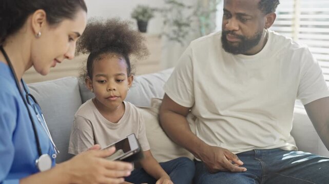 Nurse shows a digital tablet to a curious young girl sitting between her and her father, explaining a health checkup in a bright family living room.