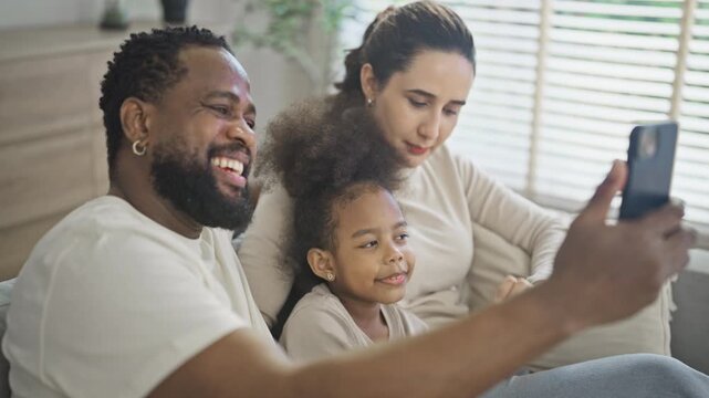 Cheerful multiracial family of three laugh together while the father holds up a smartphone to take a family selfie on the living room sofa at home in soft daylight.
