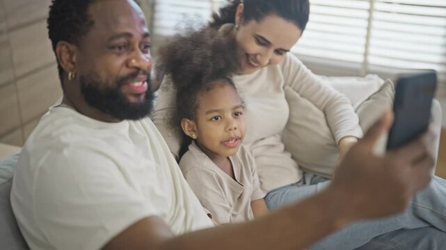 Multiracial family of three wave and smile at a smartphone during a video call with relatives, sitting close together on the living room sofa in a bright home.