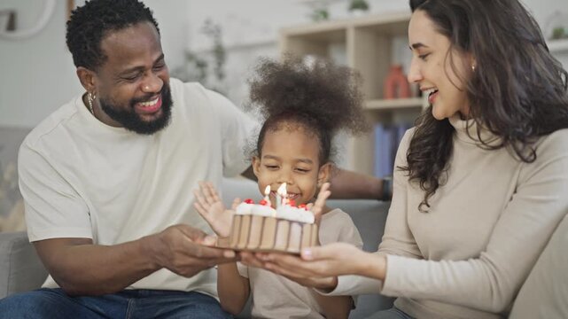Excited young girl leans in to blow out the candles on her birthday cake while her smiling multiracial parents hold the cake steady in a bright home living room.