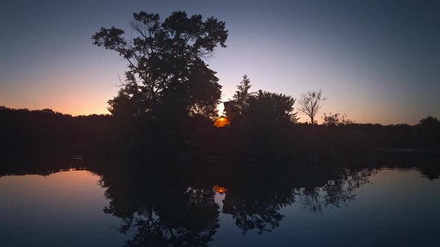 Glowing tent among trees on a small island in a calm lake at dusk in Ukraine. Tourists camping after SUP paddleboarding across the water during twilight.