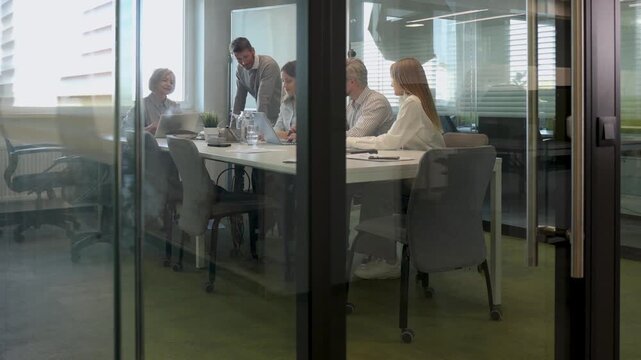 Group of business people standing together and discussing their work and projects, having a team meeting in an office, view from glass wall