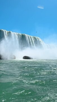 Waterfall cascading into turbulent river water. White foam covers green river surface near misty base of tall rushing waterfall.
