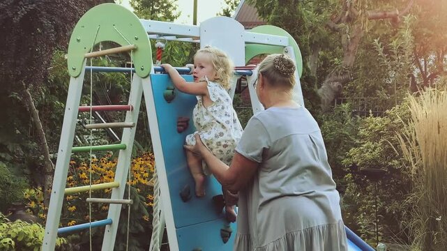 Grandmother helps toddler granddaughter climb an outdoor playground wall, supportive grandparenting, active childhood, family bonding and summer play.