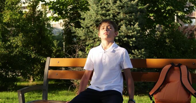 Schoolboy sits on park bench with backpack. Student rests in warm sunlight and enjoys summer nature. Kid pauses outdoors after school, showing peaceful youth lifestyle and mindful relaxation.