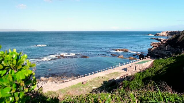 A cinematic drone shot reveals scenic coastal beauty, with a natural tidal pool reflecting the clear blue sky, dynamic waves crashing, and lush green foliage framing the view.