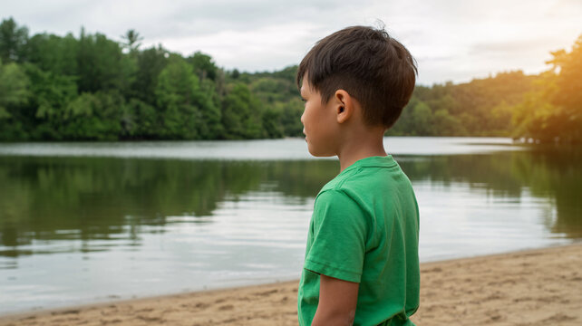 Pensive young boy standing by a calm lake, thoughtful child looking at water, peaceful summer nature scene at a sandy beach
