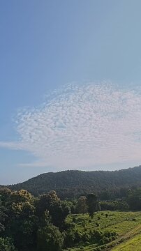 Timelapse of textured white clouds moving across a blue sky above a forested mountain and green meadow, capturing peaceful tropical nature and scenic outdoor landscape.
