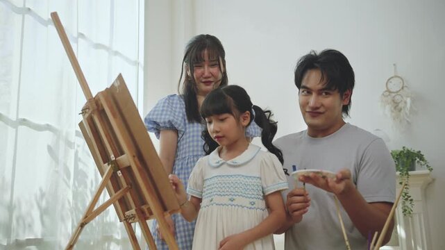 Asian family enjoying painting together at home. father, mother, and young daughter doing creative art activity with paint and canvas in a bright living room, creativity, and learning through play.