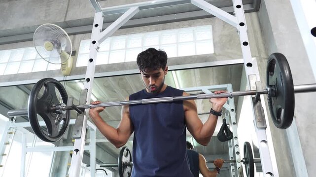 Asian man performing a barbell back squat exercise inside a power rack. He looking straight ahead with concentration. Ideal for health, fitness, weight training, bodybuilding, and active lifestyle.