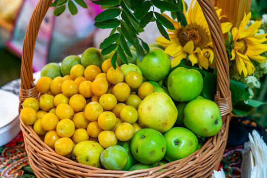 Wicker basket filled with yellow mirabelle plums and green apples decorated with yellow sunflowers and rowan branch close-up view in bright daylight.