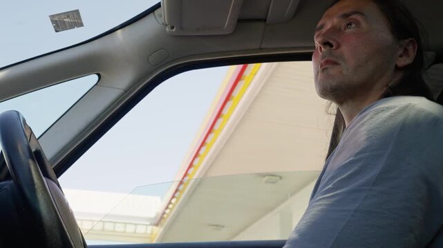 Low-angle interior shot of male driver holding steering wheel beneath gas station canopy, with driver, man, car, interior detail, showing steering, wheel, hand, drive context