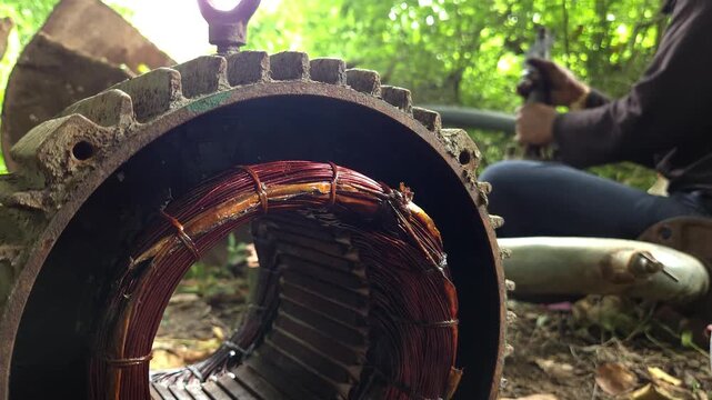 Close Up Detail of Copper Wound Stator Tunnel with an Industrial Mechanic Polishing a Metal Shaft and Removing Rust with Sandpaper in the Background of an Outdoor Machinery Maintenance Setting